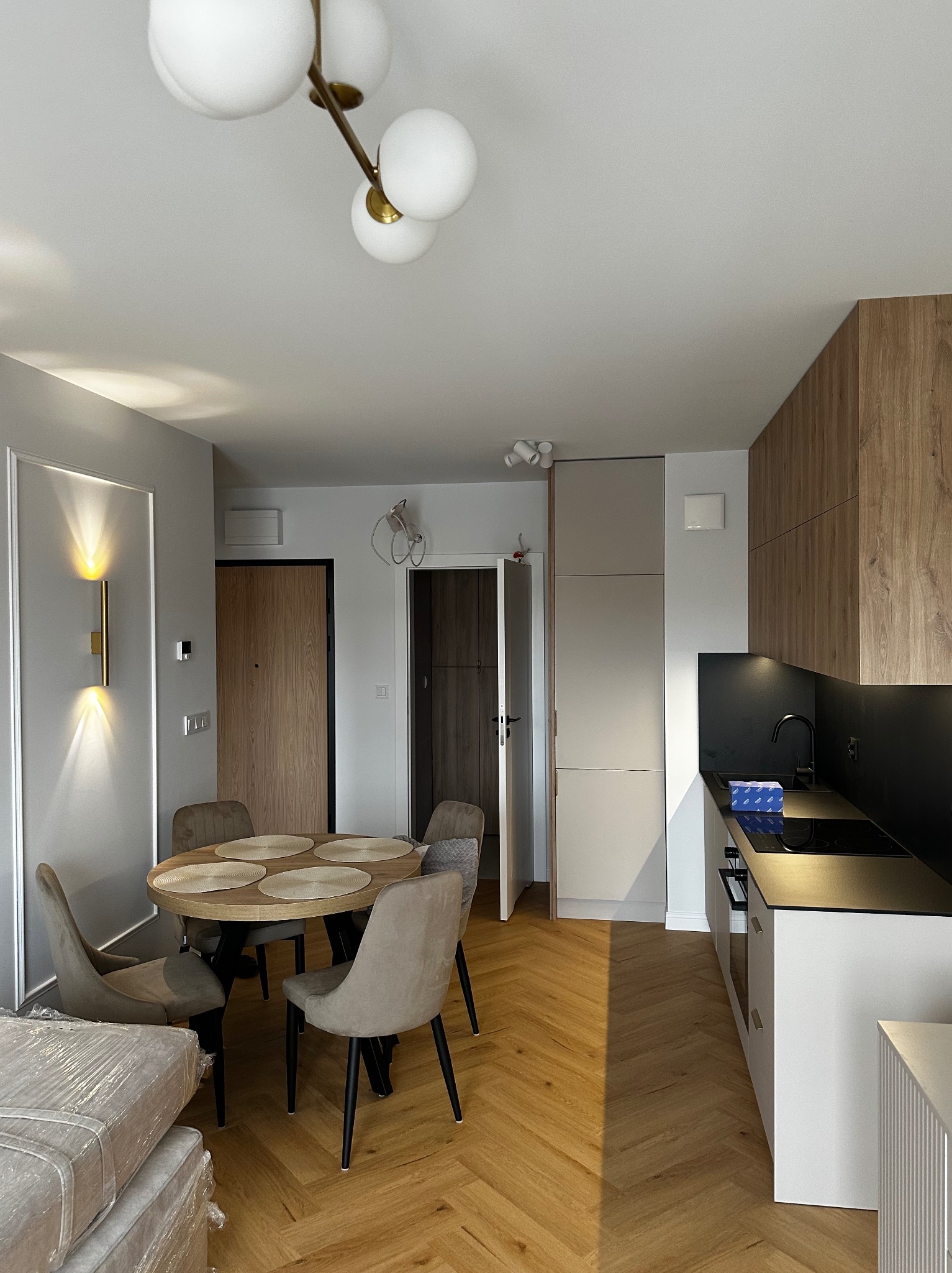 Jasne wnętrze mieszkania z parkietem w jodełkę, okrągłym stołem i nowoczesną zabudową kuchenną z drewnianymi frontami. Widoczne oświetlenie i elementy dekoracyjne.