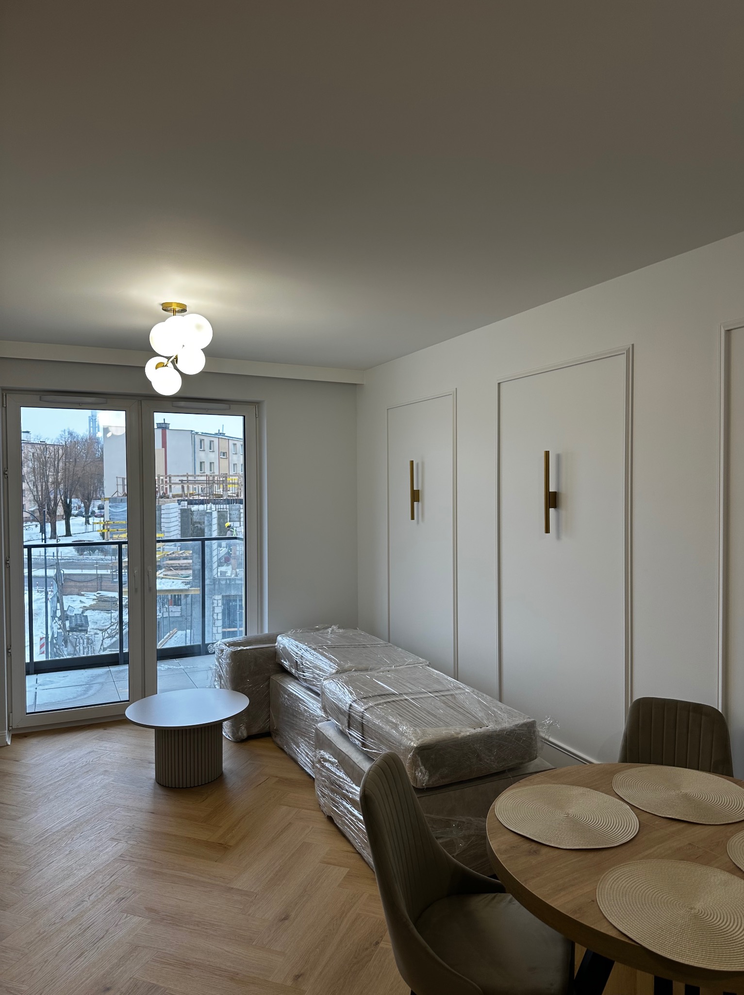 Jasne wnętrze po remoncie: parkiet w jodełkę, białe ściany z dekoracyjnymi listwami, nowoczesne lampy ścienne i sufitowa, meble zabezpieczone folią, widok na budowę z balkonu.