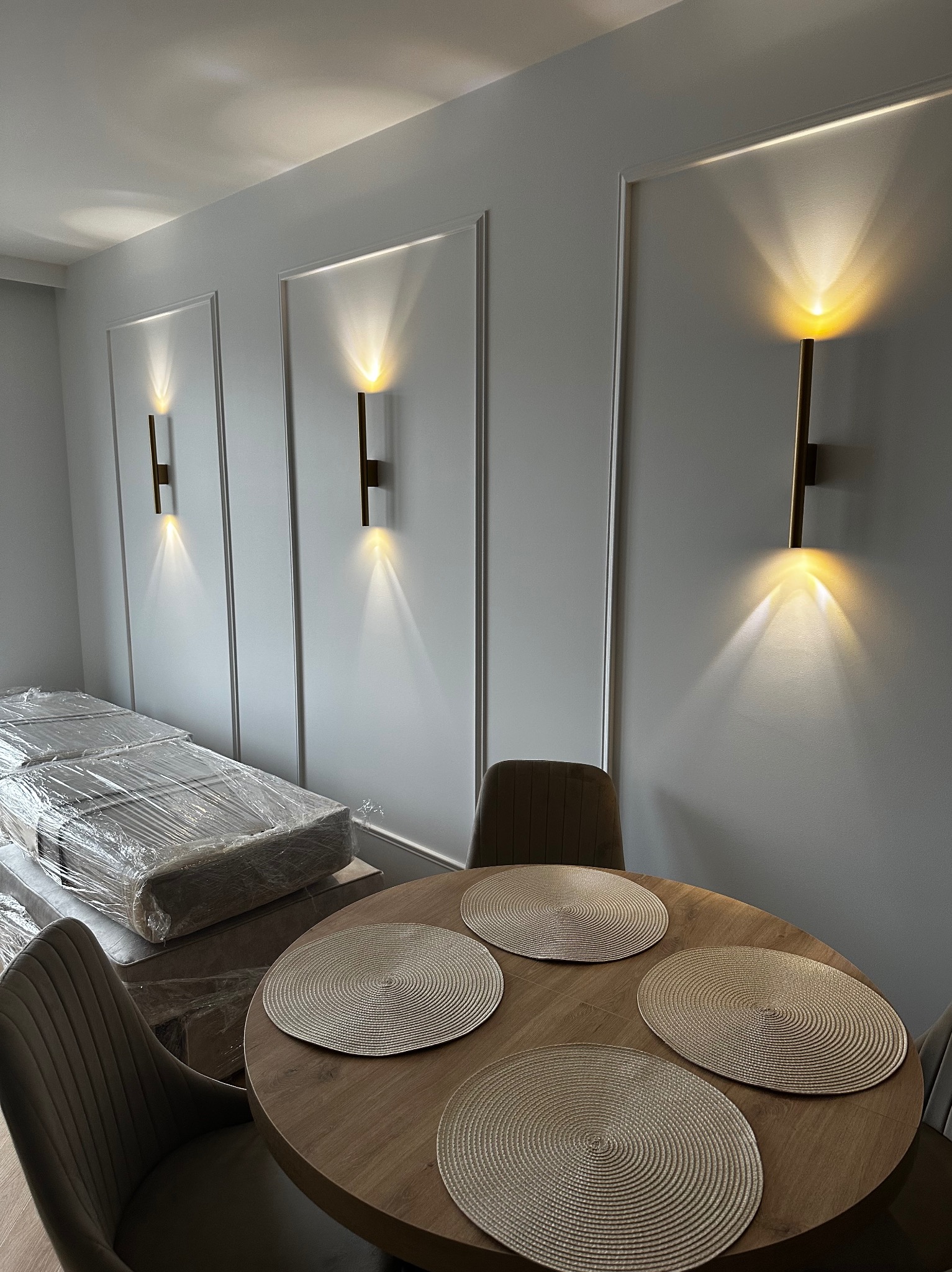 Szare ściany z dekoracyjnymi listwami i nowoczesnymi kinkietami nad okrągłym stołem z podkładkami. W tle meble zabezpieczone folią, krzesła w kolorze taupe.