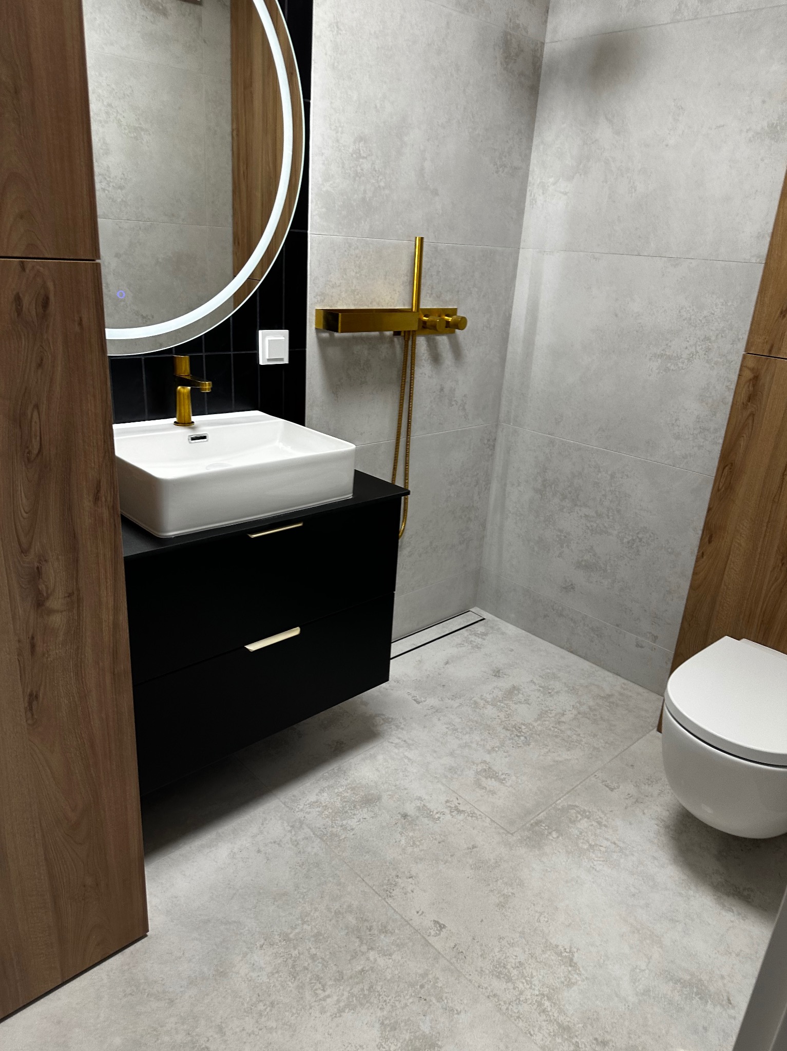 Nowoczesna łazienka z umywalką nablatową, złotą armaturą i podświetlanym lustrem. Szare płytki imitujące kamień na ścianach i podłodze. Minimalistyczny design.