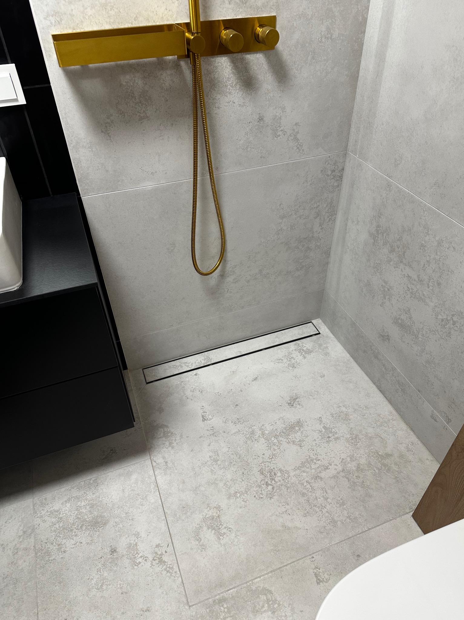 Nowoczesna kabina prysznicowa z odpływem liniowym i złotą armaturą na tle szarych, teksturowanych płytek. Minimalistyczny design, eleganckie wykończenie.