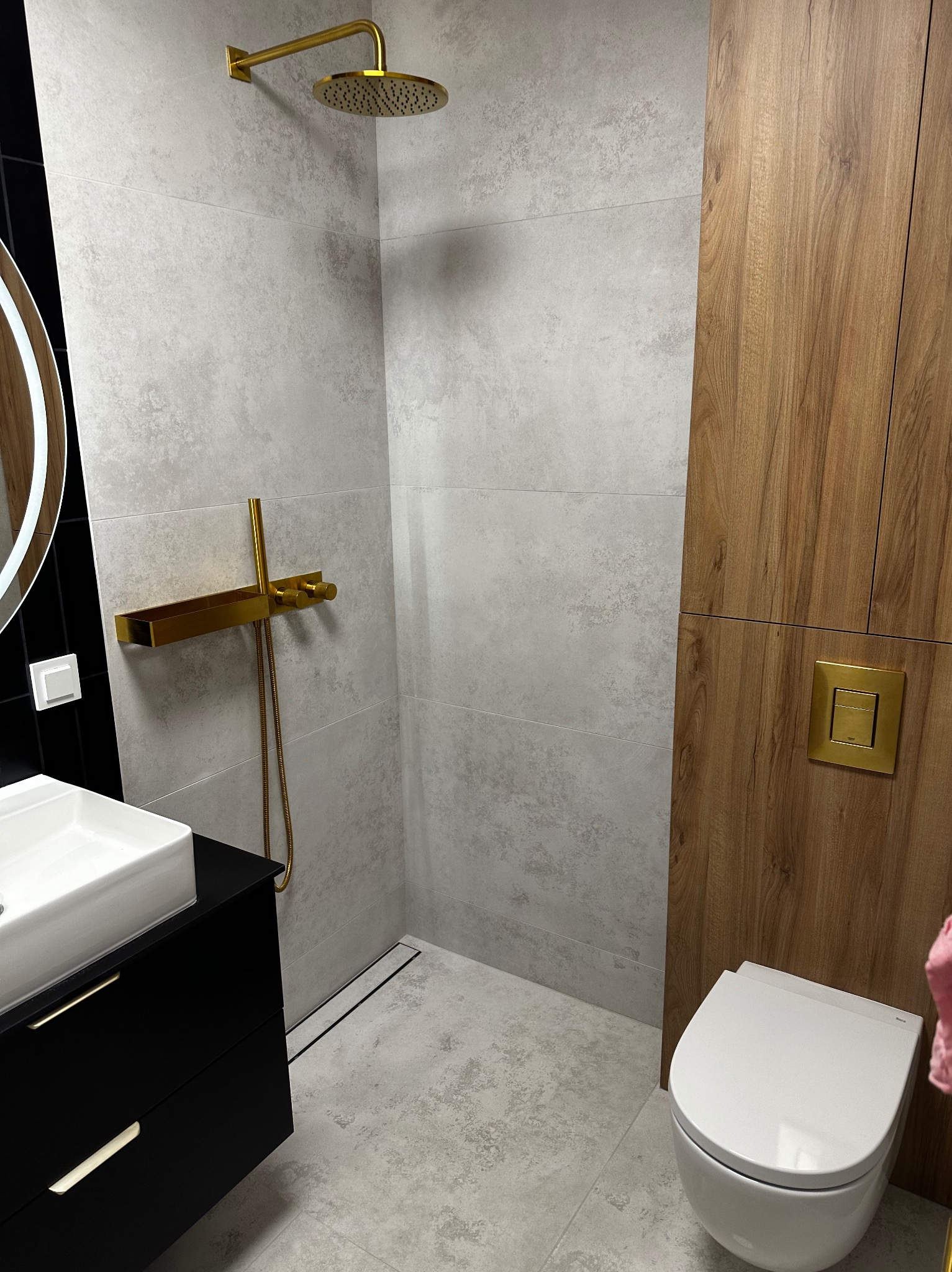 Nowoczesna łazienka z kabiną prysznicową typu walk-in, armaturą w kolorze złotym, umywalką z czarną szafką i drewnianą zabudową. Minimalistyczny design.