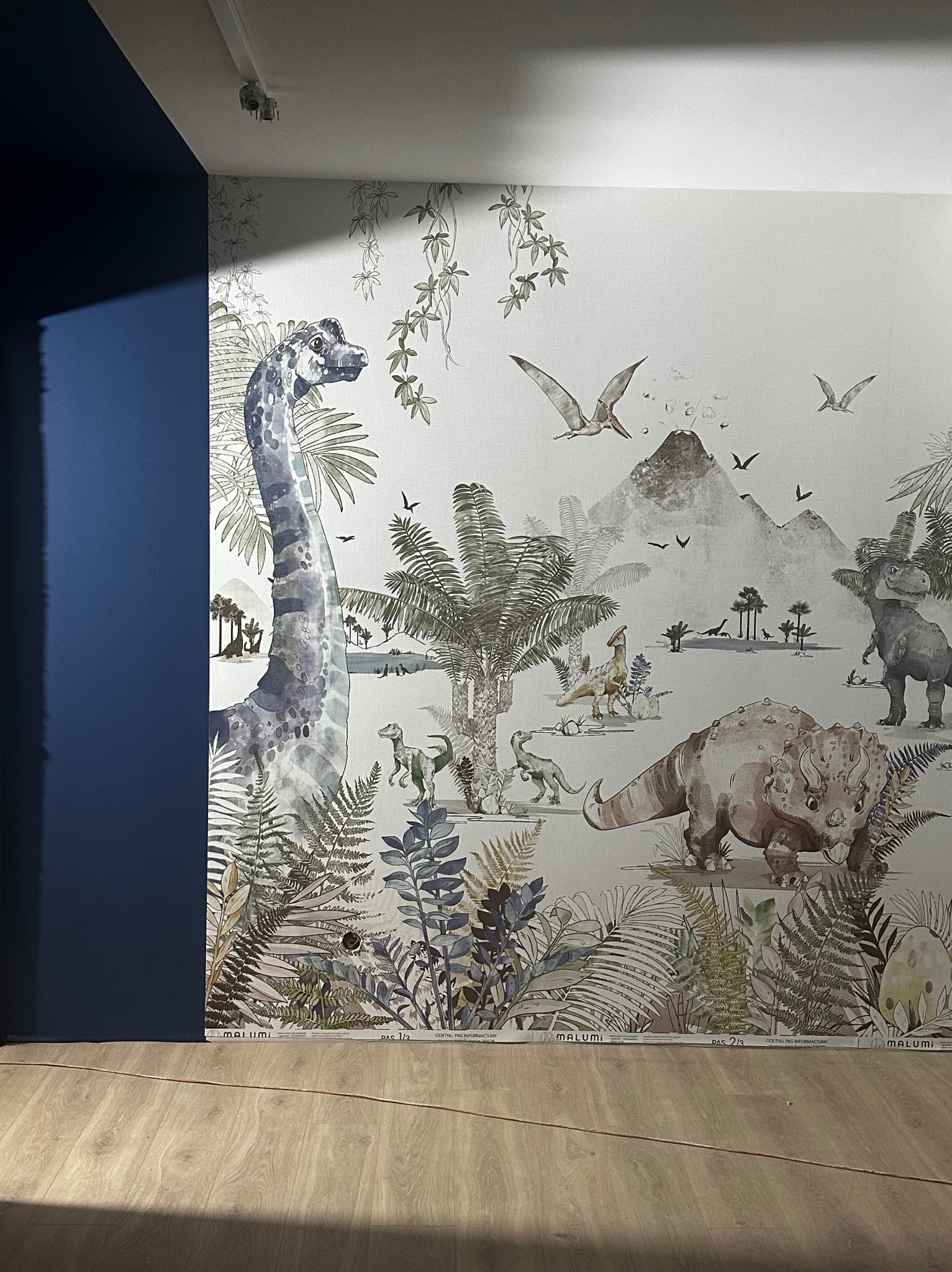 Ściana z tapetą w motywy prehistoryczne: dinozaury, roślinność, wulkan. Fragment niebieskiej ściany po lewej. Widoczna podłoga i listwa przypodłogowa z nazwą producenta tapety.