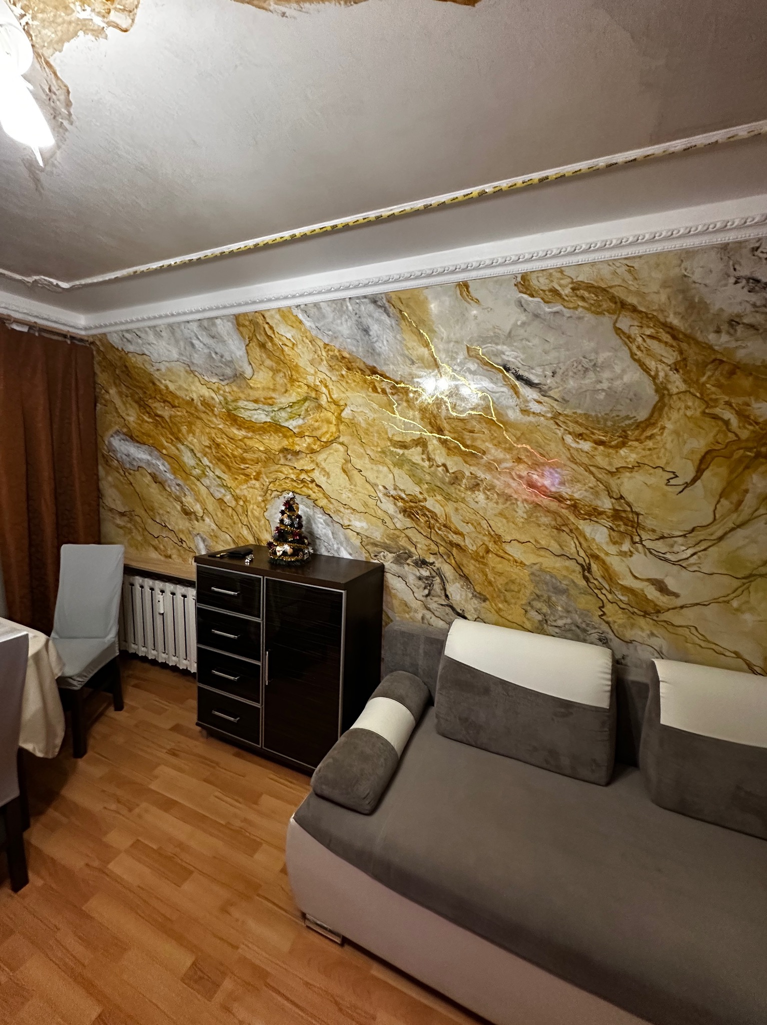 Salon po remoncie z dekoracyjną ścianą w odcieniach złota i szarości, ciemna komoda z choinką, szara sofa i drewniana podłoga. Widoczne ślady uszkodzeń na suficie.