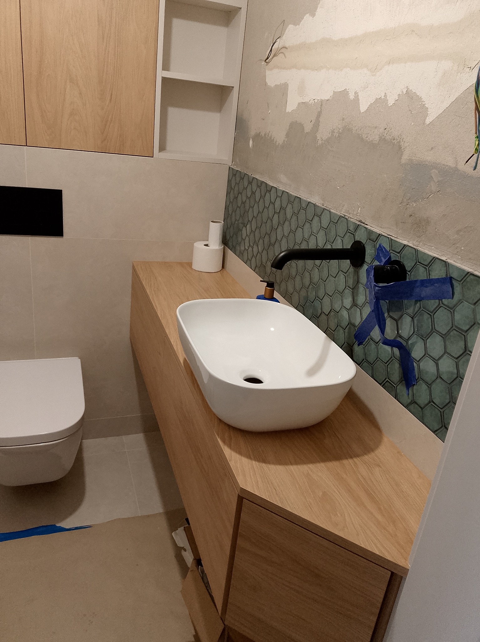 Nowoczesna łazienka w trakcie wykończenia: biała umywalka nablatowa, drewniana szafka, heksagonalne płytki na ścianie, widoczne instalacje i zabezpieczenia.