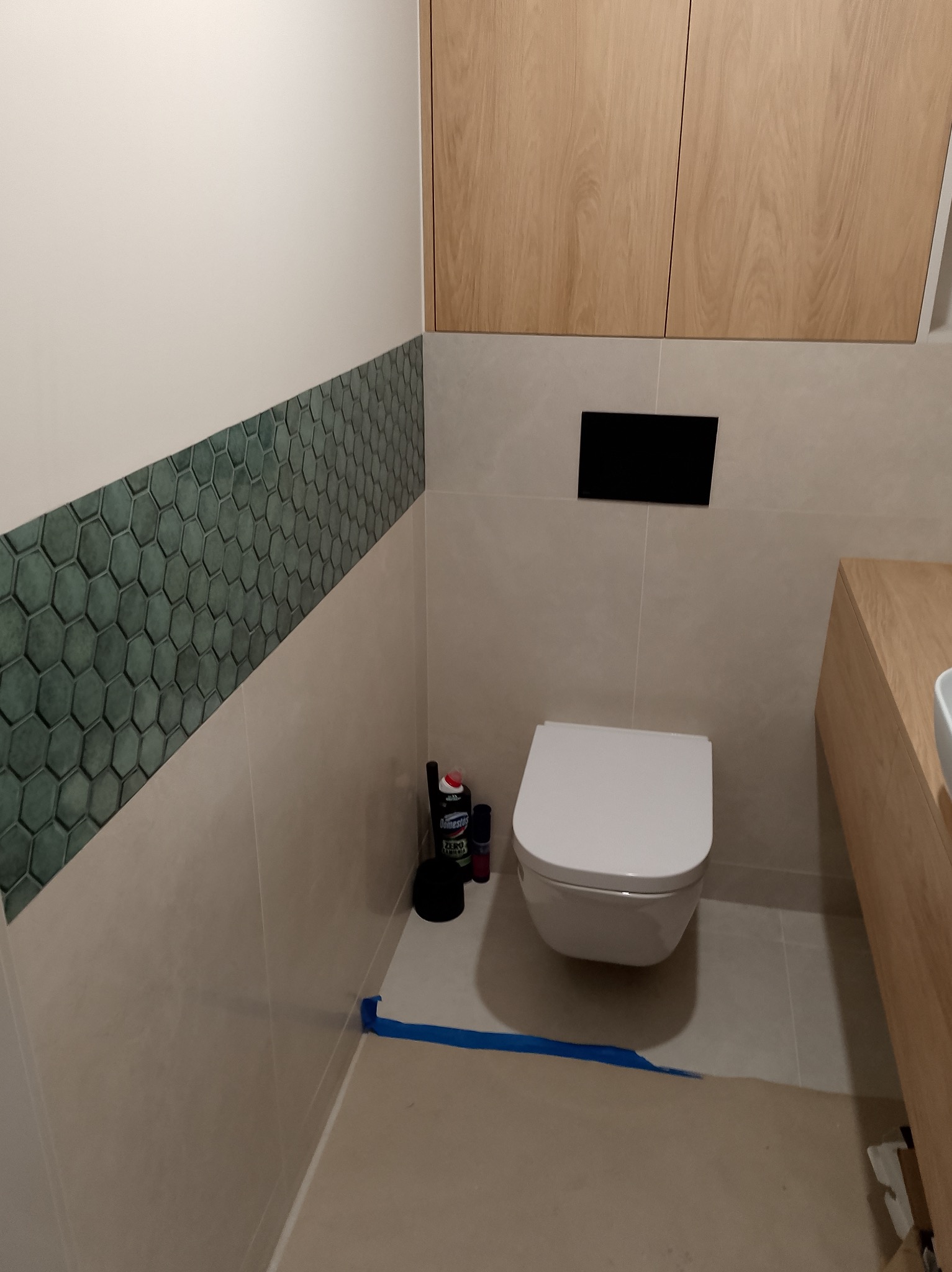 Nowoczesna łazienka z podwieszaną toaletą, heksagonalnymi płytkami i drewnianymi szafkami. Widoczne akcesoria i taśma malarska, sugerujące świeżo zakończony remont.