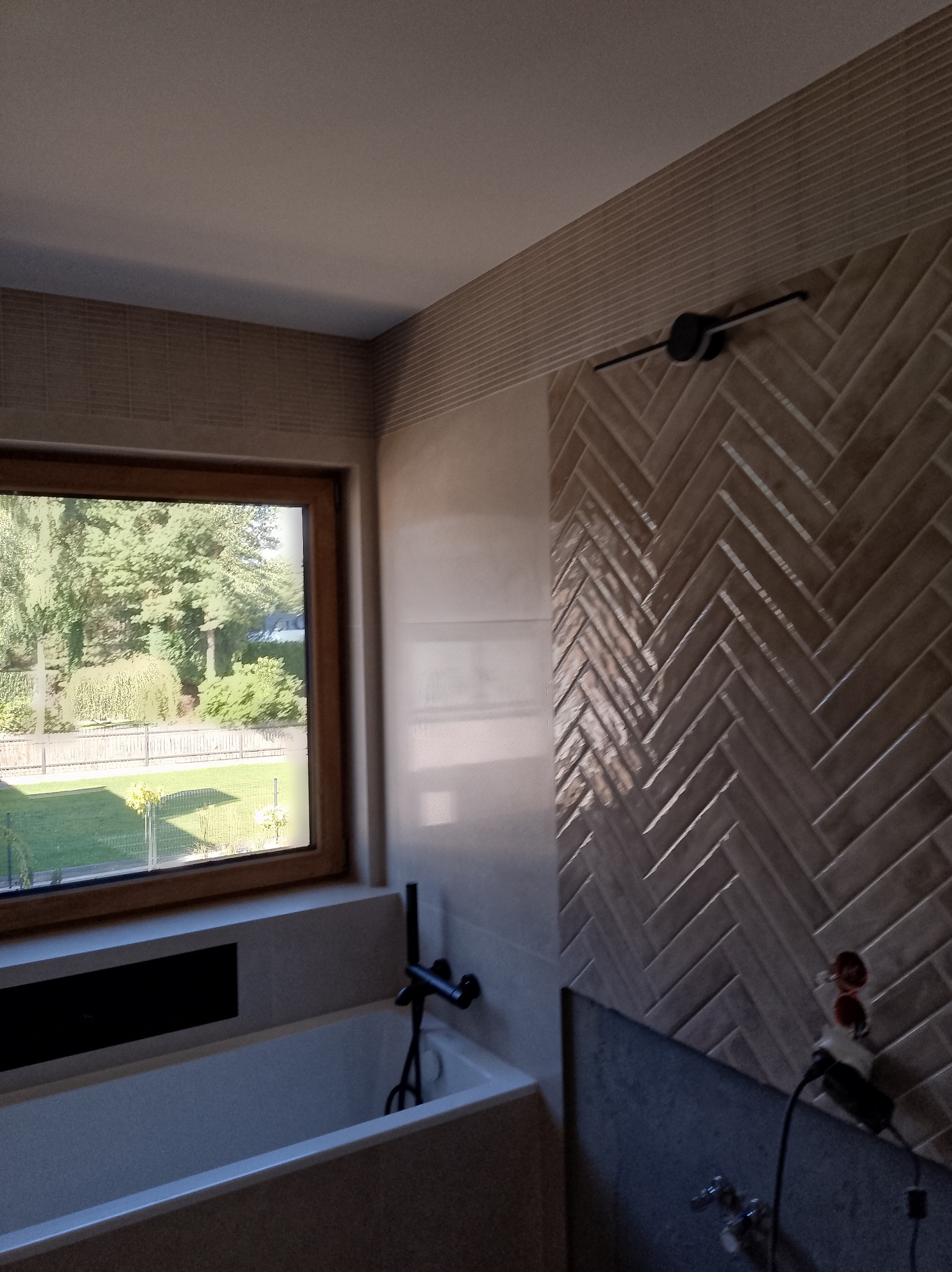 Wykończona łazienka z wanną, oknem z widokiem na zieleń i ścianą z płytek ułożonych w jodełkę. Widoczne elementy armatury i fragment instalacji elektrycznej.