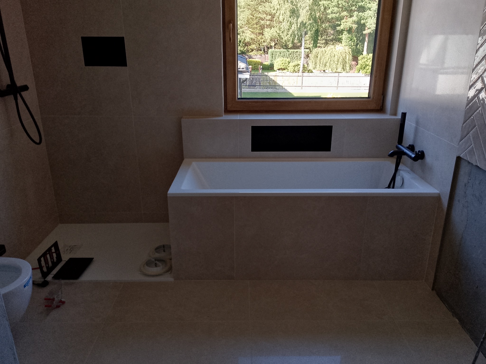 Wykończona łazienka z beżowymi płytkami, białą wanną i czarną armaturą. Widok na okno z zielenią na zewnątrz. Częściowo widoczna toaleta i narzędzia.