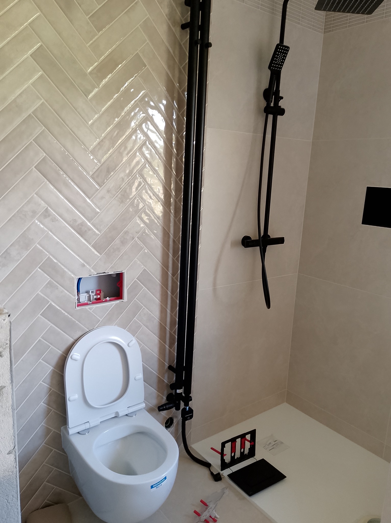 Nowoczesna łazienka w trakcie wykańczania: biała toaleta z otwartą klapą, czarna armatura prysznicowa, ściany wyłożone beżowymi płytkami w jodełkę.