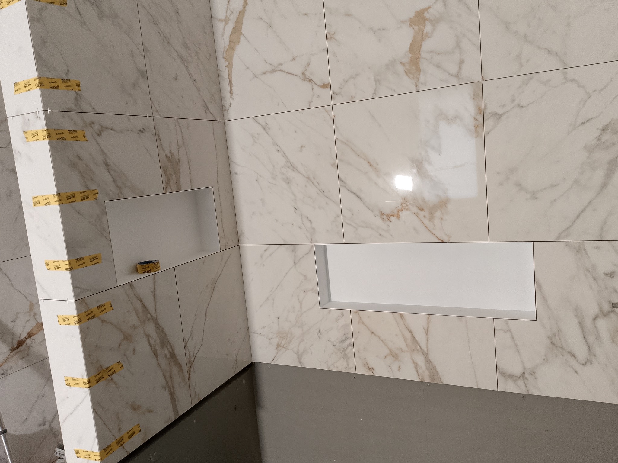 Białe płytki imitujące marmur z wnękami w trakcie montażu. Widoczne taśmy maskujące i krzyżyki dystansowe. Szara podłoga w tle.