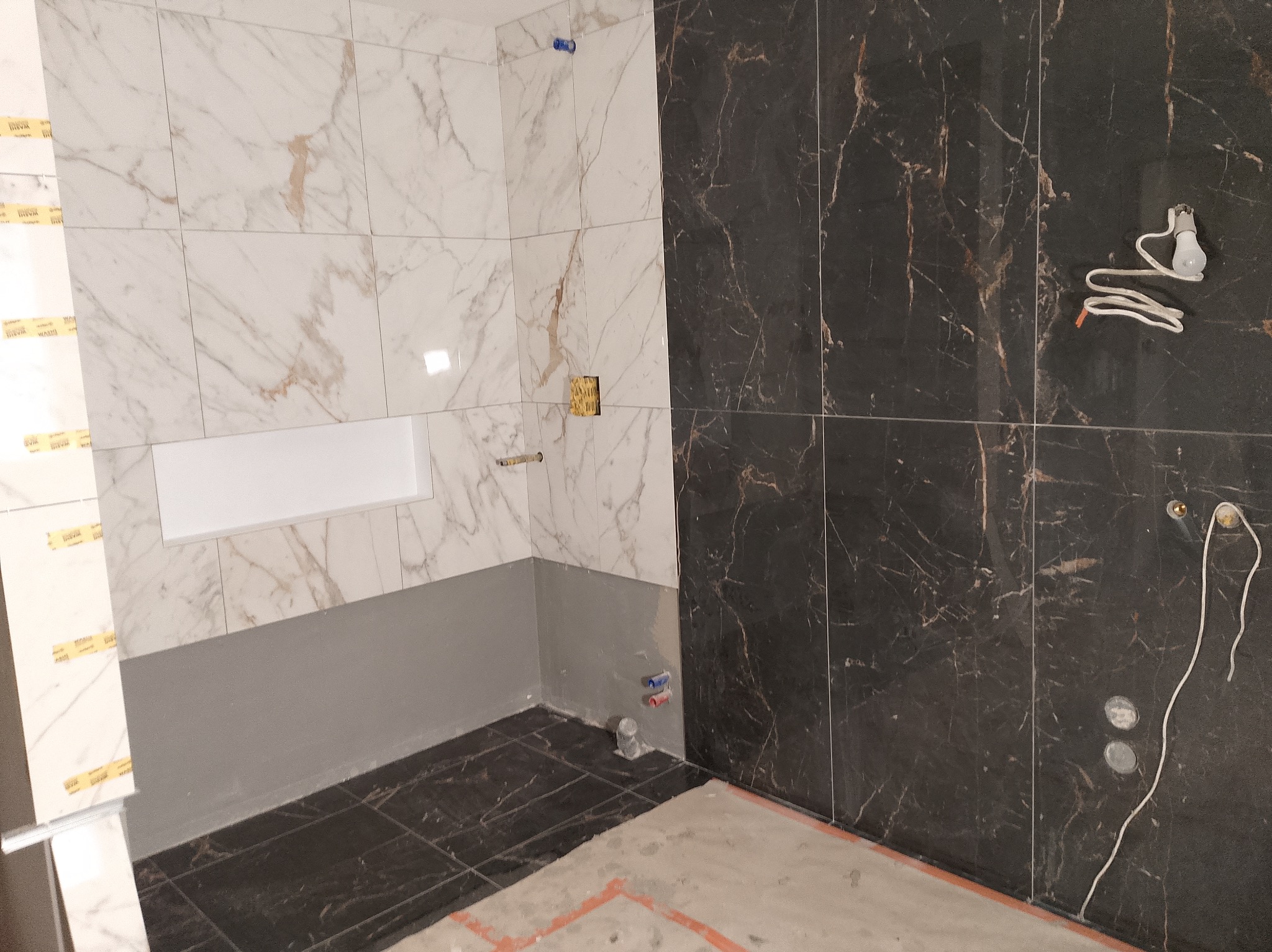 Częściowo wykończona łazienka z czarno-białymi marmurkowymi płytkami, szarą hydroizolacją i widocznymi instalacjami elektrycznymi i hydraulicznymi.