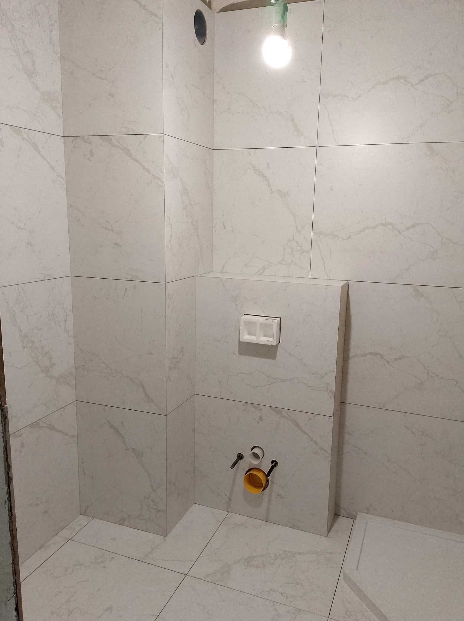 Wykończona łazienka z białymi płytkami imitującymi marmur, widoczne przyłącza do toalety podtynkowej i fragment brodzika. Oświetlenie tymczasowe.