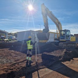 ANKUS - Żółta koparka New Holland na placu budowy, pracownik z łatą niwelacyjną, wykop pod fundamenty, błękitne niebo z promieniami słońca.