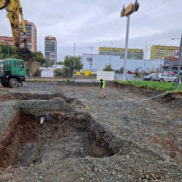 ANKUS - Wykop pod fundamenty z widoczną koparką New Holland i ciężarówką na tle zabudowy miejskiej. Pracownik w żółtej kamizelce nadzoruje prace ziemne.