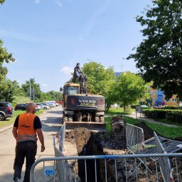 ANKUS - Żółta koparka Volvo Ankush na tle wykopu zabezpieczonego barierkami, pracownik w kamizelce idzie chodnikiem. Błękitne niebo i zieleń drzew w tle.