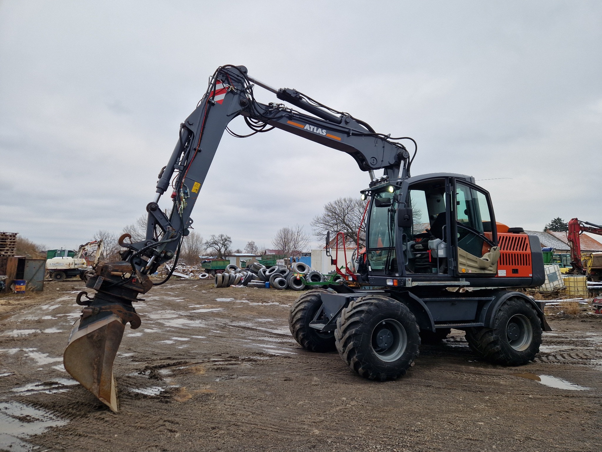 Szara koparka kołowa Atlas na błotnistym placu budowy, z podniesionym ramieniem i łyżką. W tle składowisko opon i inne maszyny budowlane. Pochmurne niebo.