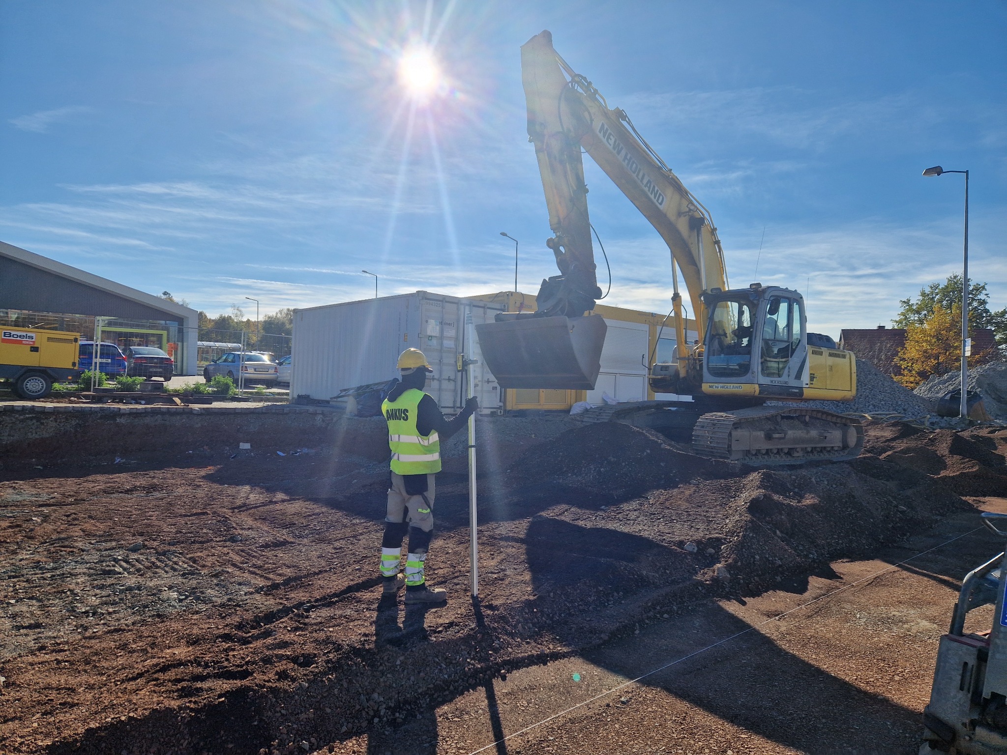 Żółta koparka New Holland na placu budowy, pracownik z łatą niwelacyjną, wykop pod fundamenty, błękitne niebo z promieniami słońca.