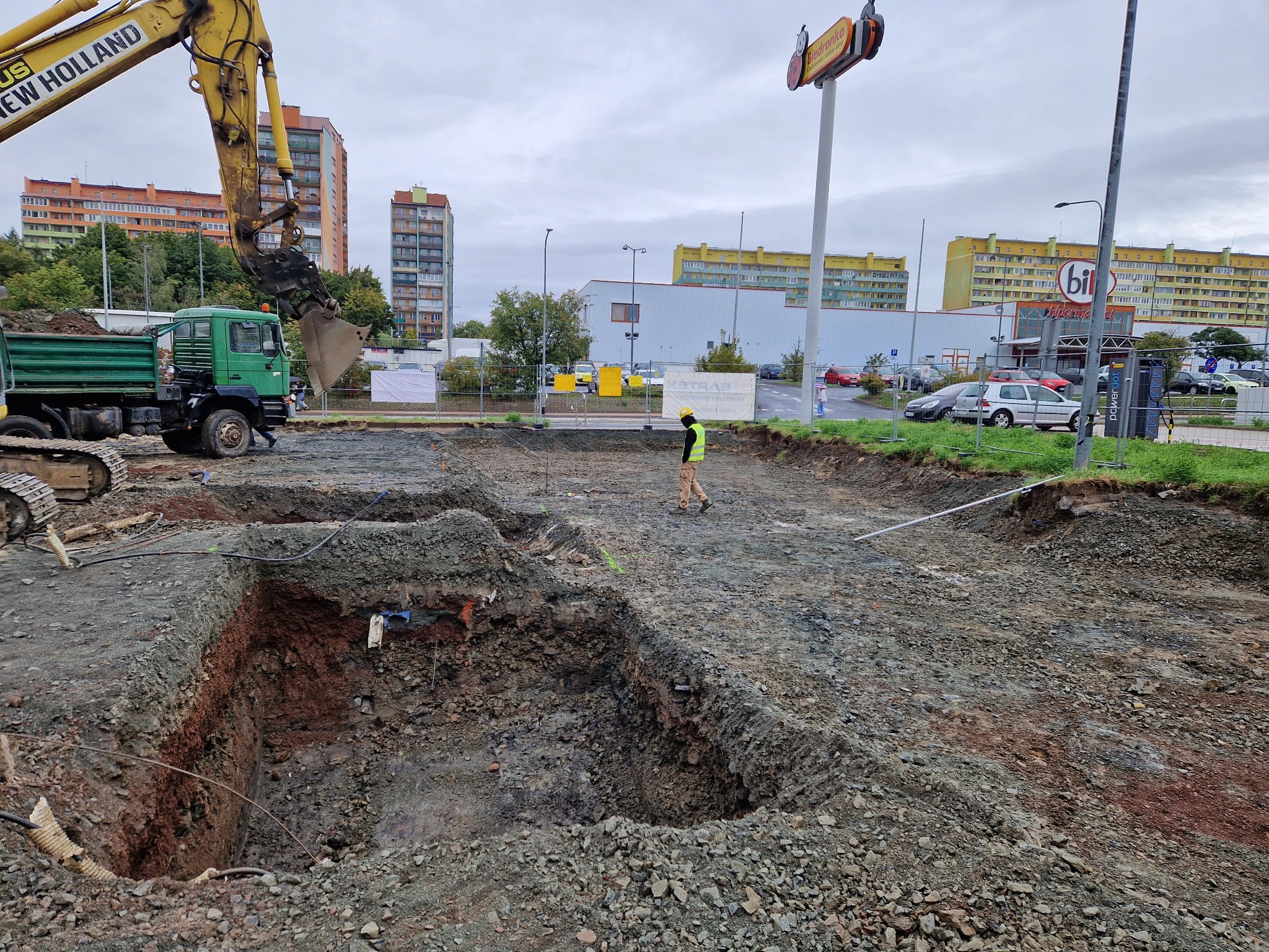 Wykop pod fundamenty z widoczną koparką New Holland i ciężarówką na tle zabudowy miejskiej. Pracownik w żółtej kamizelce nadzoruje prace ziemne.