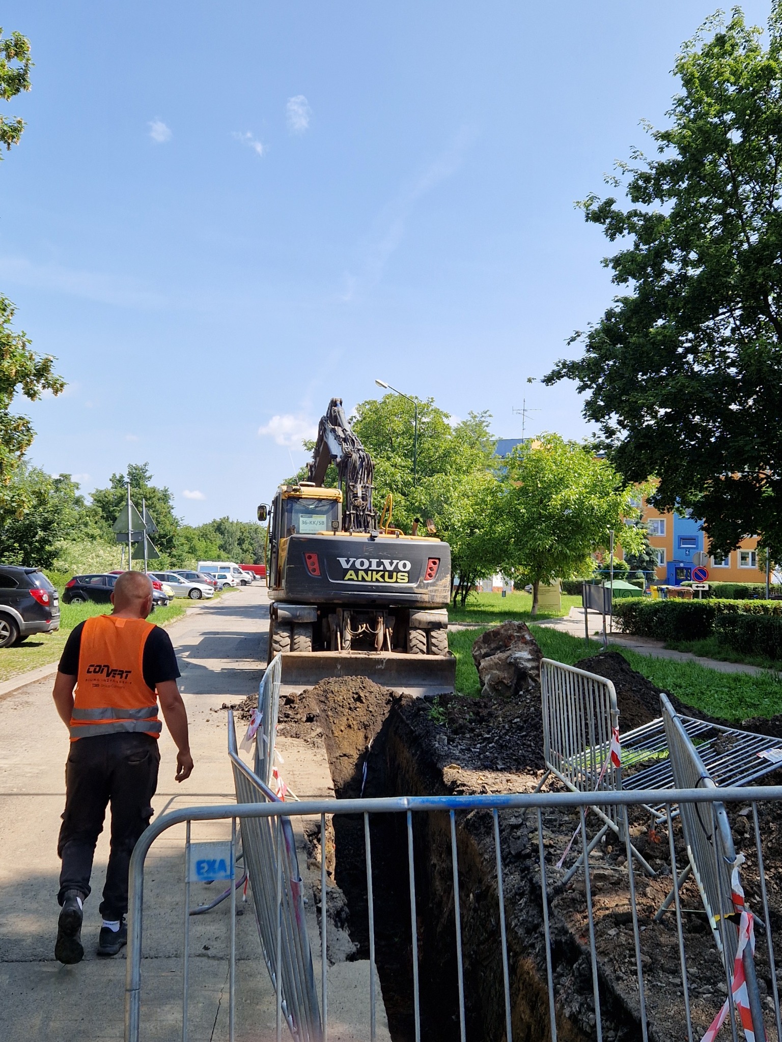 Żółta koparka Volvo Ankush na tle wykopu zabezpieczonego barierkami, pracownik w kamizelce idzie chodnikiem. Błękitne niebo i zieleń drzew w tle.