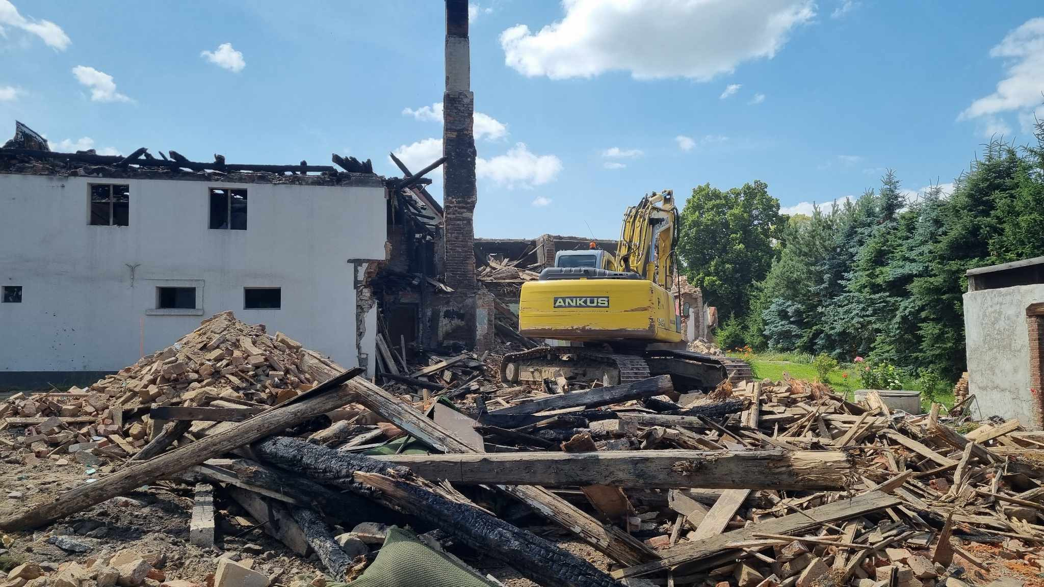 Żółta koparka ANKUS na tle częściowo zburzonego budynku z ceglanym kominem, gruz i drewniane elementy na pierwszym planie, błękitne niebo z chmurami w tle.