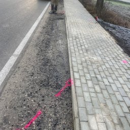 Vasyl Yaskovets - Świeżo ułożony chodnik z kostki brukowej, obok droga z zaparkowanym autem i pracownik w odblaskowej kamizelce. Wyznaczone różową farbą linie i punkty.