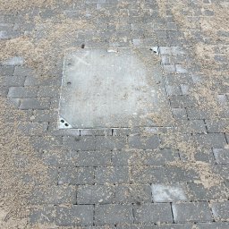 Vasyl Yaskovets - Szara kostka brukowa z piaskiem i włazem kanalizacyjnym. Widok z góry, ukończona praca brukarska. Właz wkomponowany w kostkę.