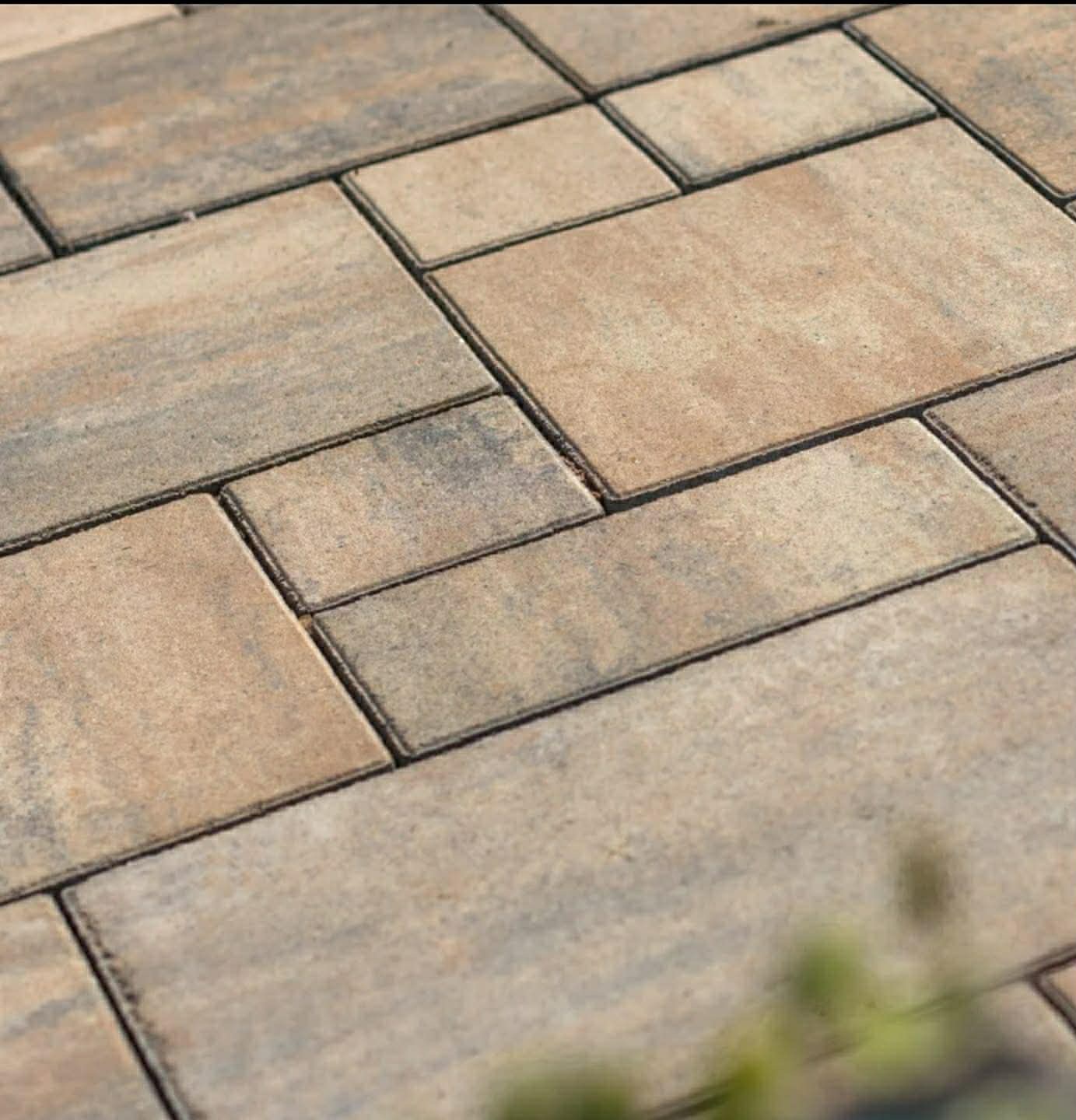 Beżowo-szara kostka brukowa ułożona w regularny wzór, widok z góry, z delikatnym rozmyciem roślinności w dolnej części kadru.