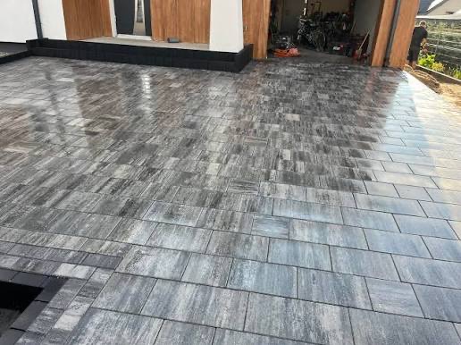 Ułożona, szara kostka brukowa na podjeździe z widocznym wejściem do garażu i domu. Kostka ułożona w regularny wzór, z widocznymi fugami.