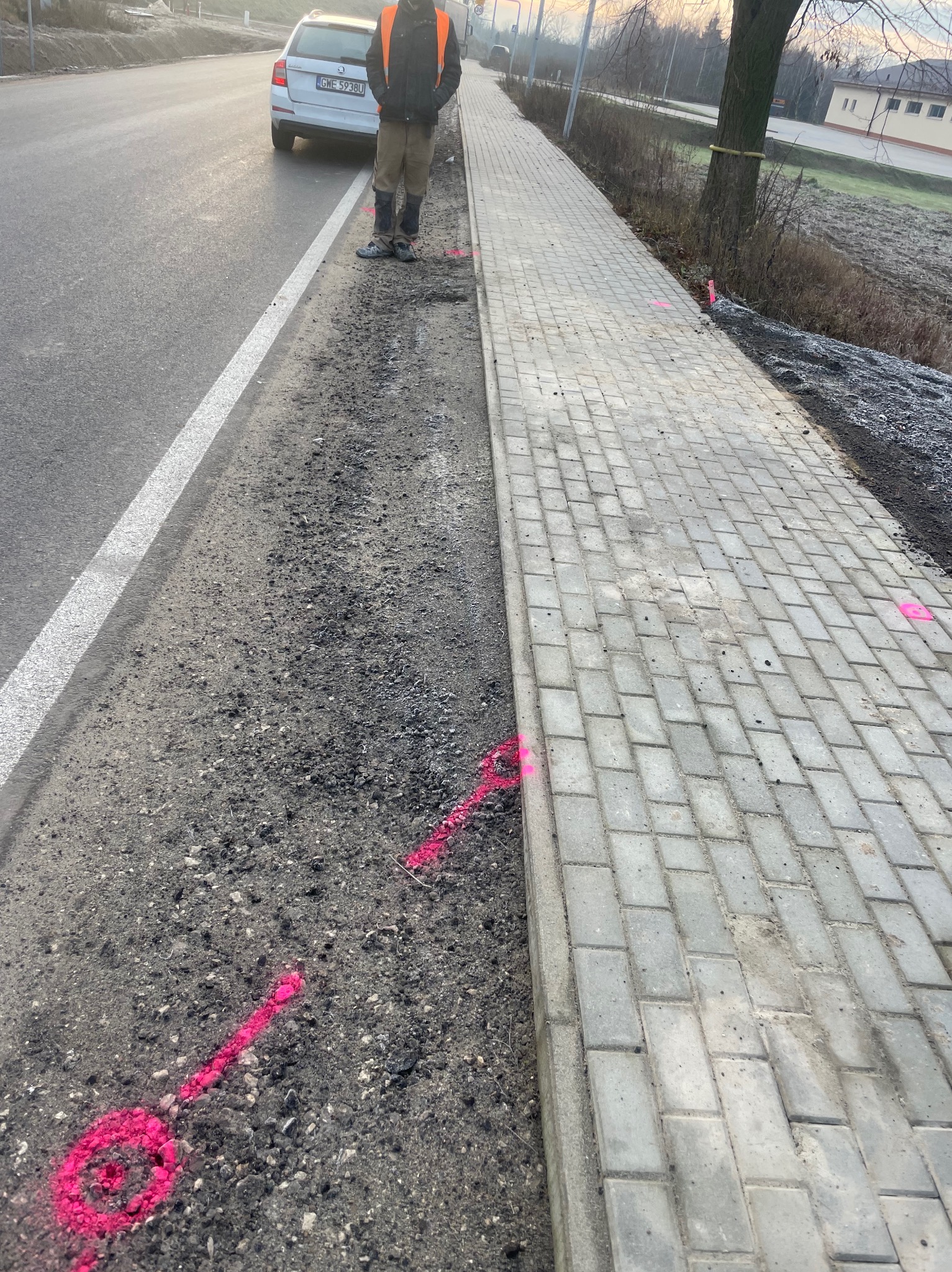 Świeżo ułożony chodnik z kostki brukowej, obok droga z zaparkowanym autem i pracownik w odblaskowej kamizelce. Wyznaczone różową farbą linie i punkty.