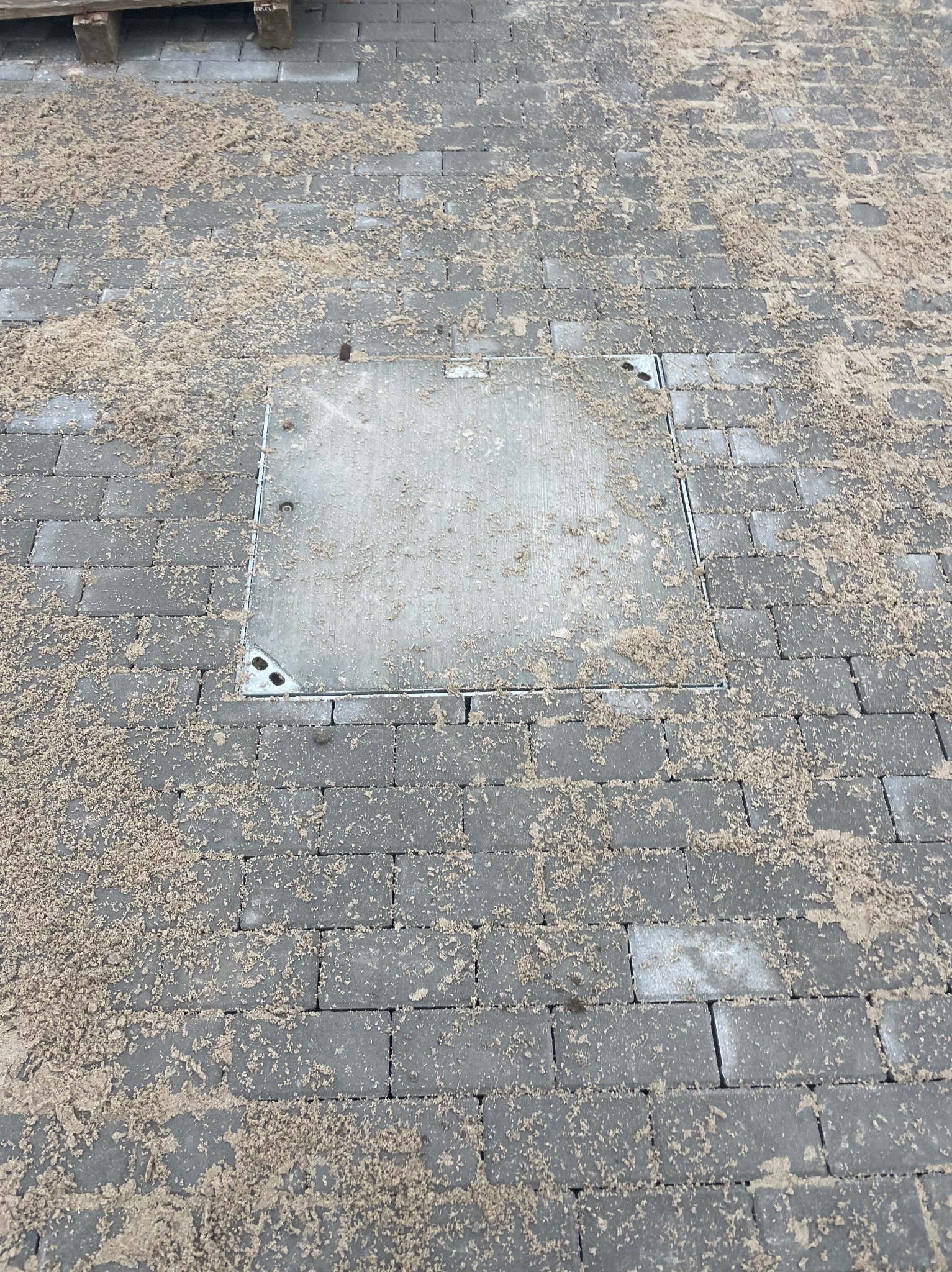 Szara kostka brukowa z piaskiem i włazem kanalizacyjnym. Widok z góry, ukończona praca brukarska. Właz wkomponowany w kostkę.