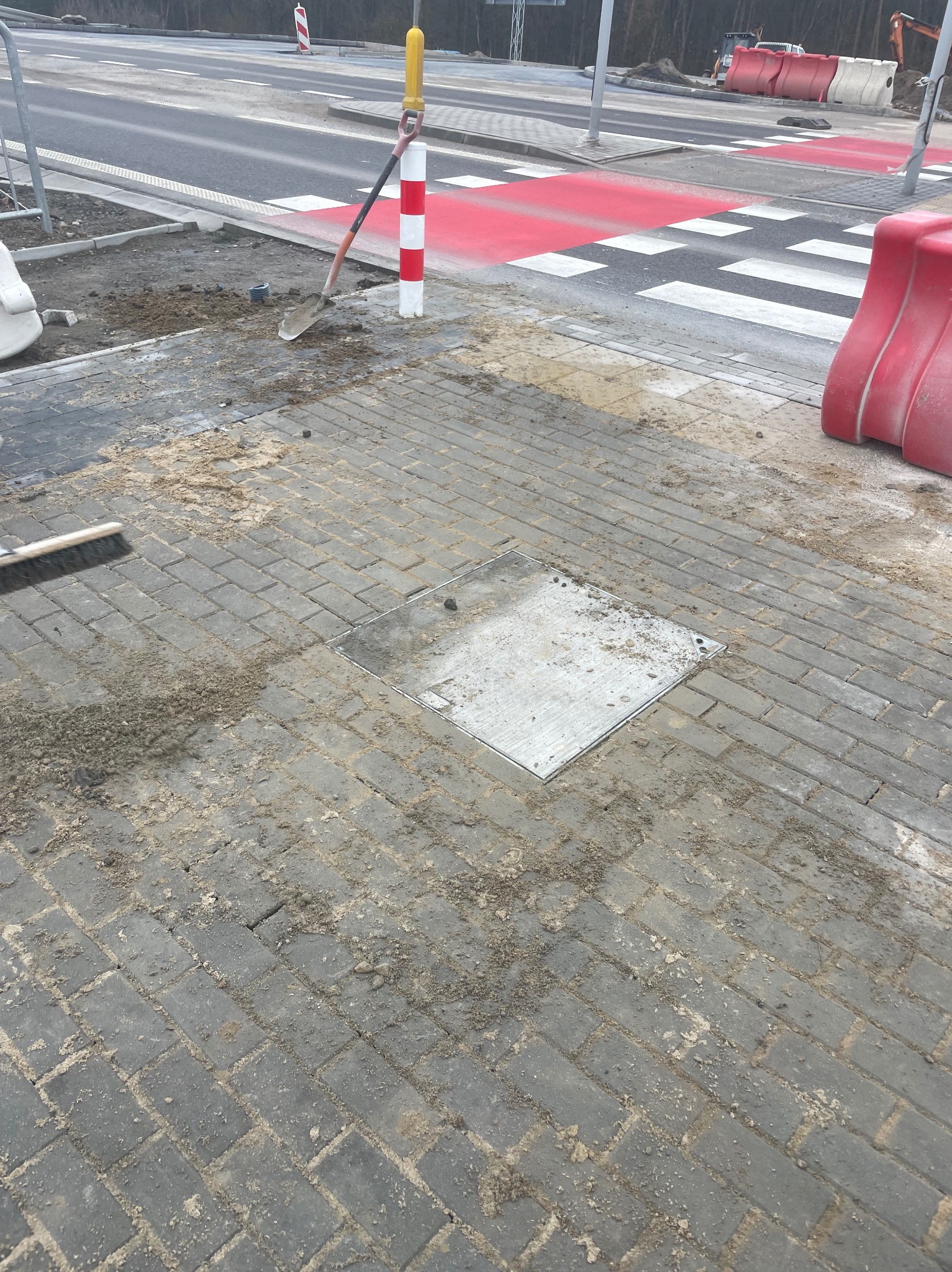 Układanie szarej kostki brukowej na chodniku przy przejściu dla pieszych. Widoczna łopata, szczotka i studzienka kanalizacyjna. Prace brukarskie w toku.