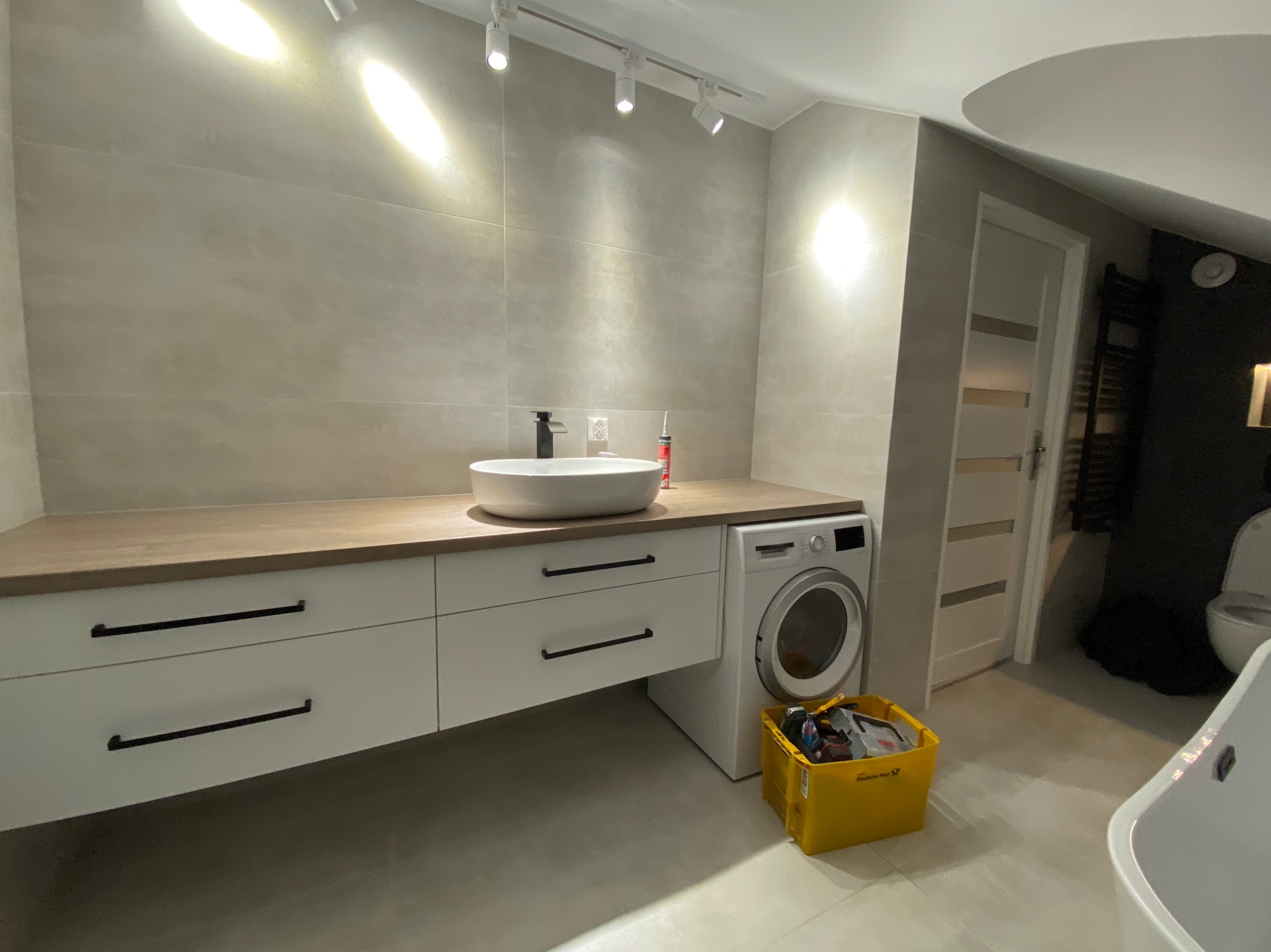 Nowoczesna łazienka z białymi szafkami pod umywalką z drewnianym blatem, pralką i żółtym koszem. Umywalka owalna, biała. Oświetlenie punktowe.