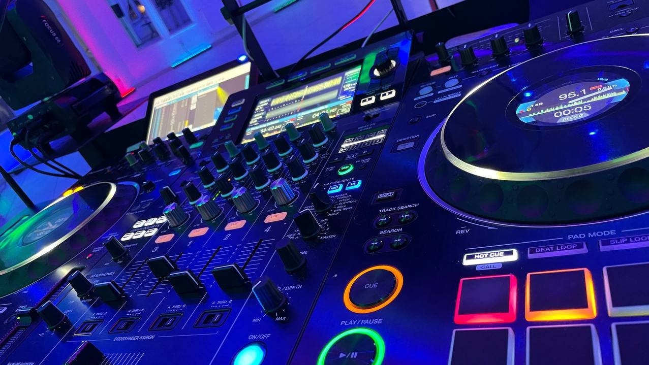 Profesjonalny kontroler DJ Pioneer z podświetleniem LED w odcieniach niebieskiego i zielonego, widoczne pokrętła, suwaki i ekran LCD z informacjami o odtwarzanym utworze.