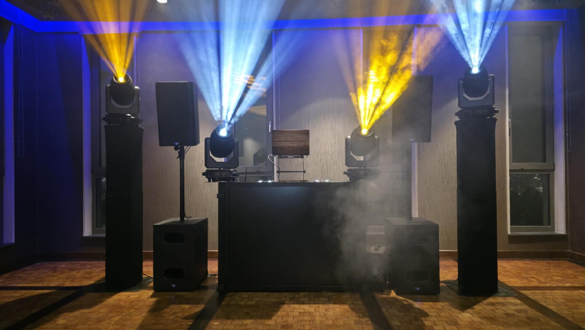 Profesjonalne nagłośnienie i oświetlenie sceniczne z reflektorami w kolorach niebieskim i żółtym, ustawione w pomieszczeniu z parkietem i oknami.