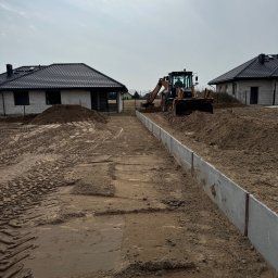 Trawtex - Prace ziemne na działce budowlanej: równanie terenu koparką przy betonowych obrzeżach. W tle dwa domy w stanie surowym pod ciemnymi dachami.