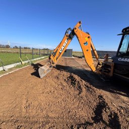 Trawtex - Pomarańczowa koparka CASE na tle przygotowanego terenu pod ogród z widocznym ogrodzeniem panelowym i turbinami wiatrowymi w oddali, w słoneczny dzień.