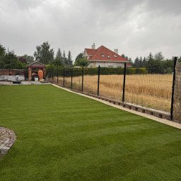 Trawtex - Nowo założony, idealnie zielony trawnik z wyraźnymi pasami, otoczony kamiennym obrzeżem i ciemnym ogrodzeniem. W tle złote pole, dom z czerwonym dachem i zachmurzone niebo.