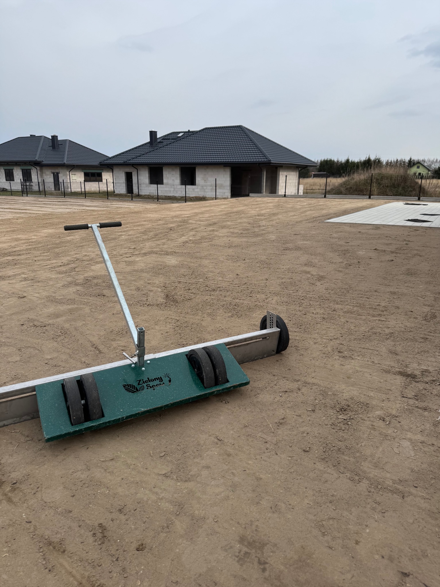 Przygotowanie terenu pod trawnik: wał z rolkami na piasku, w tle domy w stanie surowym i fragment ułożonej kostki. Sprzęt firmy Zielony Spac.