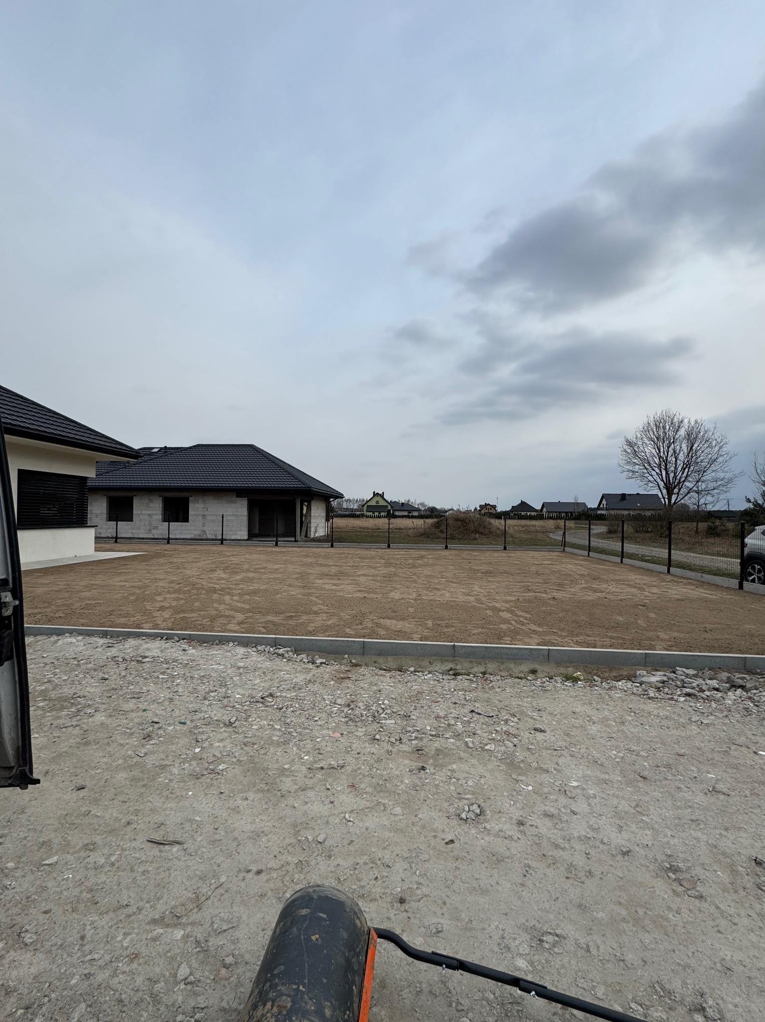 Przygotowany teren pod trawnik, widoczne ogrodzenie panelowe i budynki w tle. Walec ogrodowy na pierwszym planie, gotowy do wyrównania podłoża.