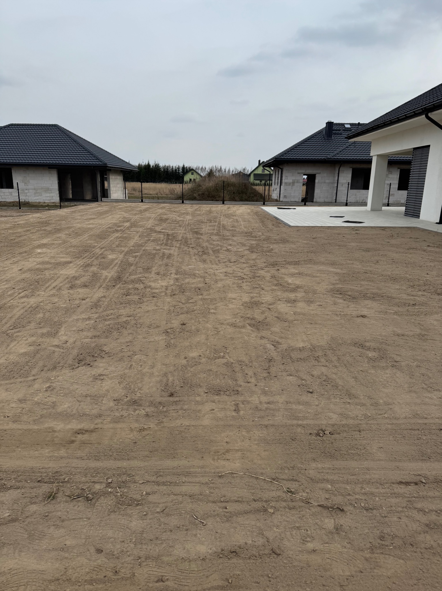 Przygotowany teren pod trawnik z widocznymi śladami opon, w tle dwa domy w stanie surowym zamkniętym i ogrodzenie z siatki.