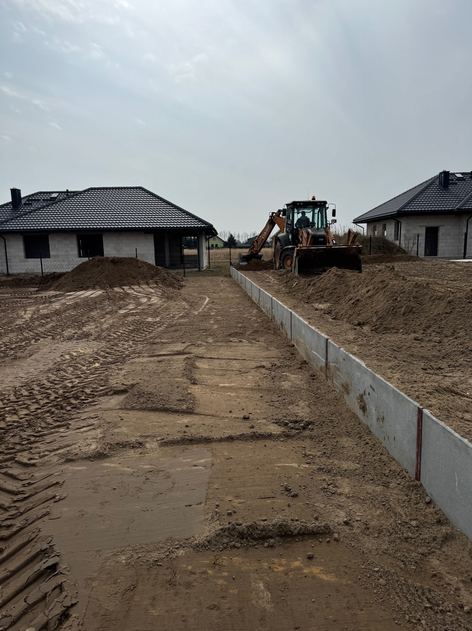 Prace ziemne na działce budowlanej: równanie terenu koparką przy betonowych obrzeżach. W tle dwa domy w stanie surowym pod ciemnymi dachami.