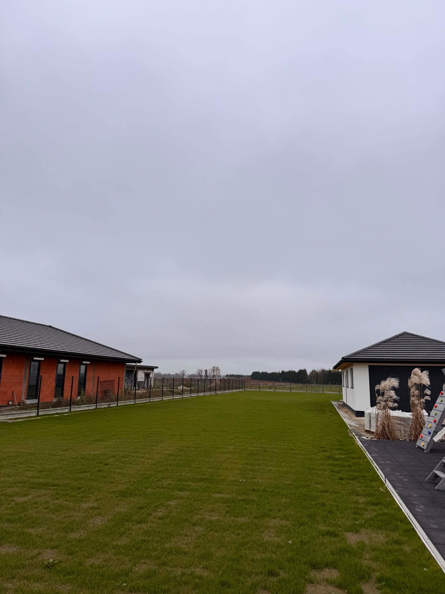 Rozległy trawnik z rolki, świeżo ułożony w Parczewie, otoczony budynkami i ogrodzeniem. Widok na posesję w pochmurny dzień.