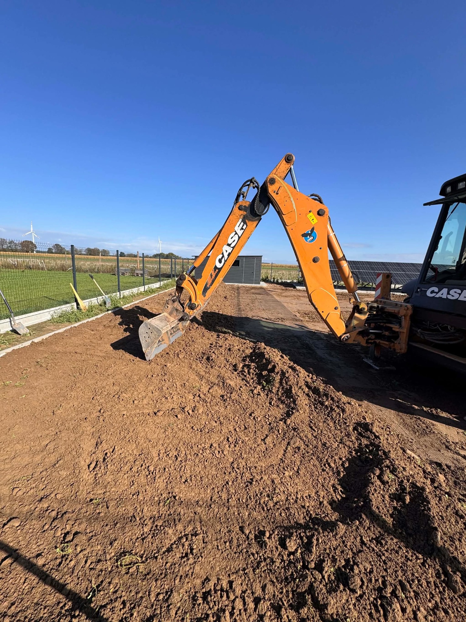Pomarańczowa koparka CASE na tle przygotowanego terenu pod ogród z widocznym ogrodzeniem panelowym i turbinami wiatrowymi w oddali, w słoneczny dzień.