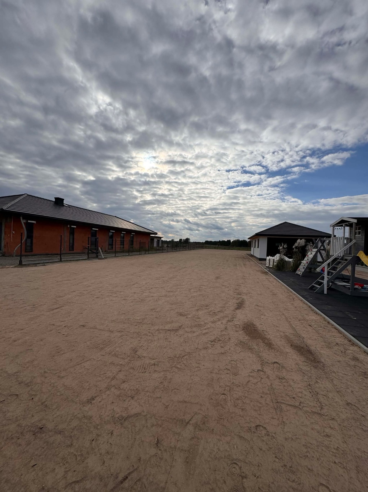 Rozległy teren z piaskowym podłożem, otoczony budynkami i ogrodzeniem. Ślady na piasku sugerują niedawne prace ziemne lub przygotowanie terenu pod inwestycję.