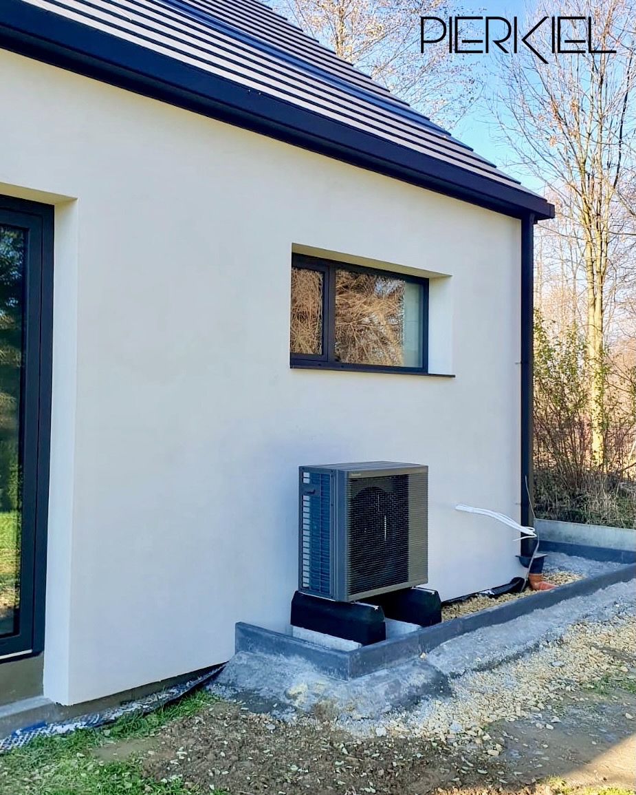 Szara pompa ciepła zamontowana na zewnątrz białego budynku z czarnymi ramami okiennymi i dachem. Widoczne elementy instalacji i otoczenie budynku.