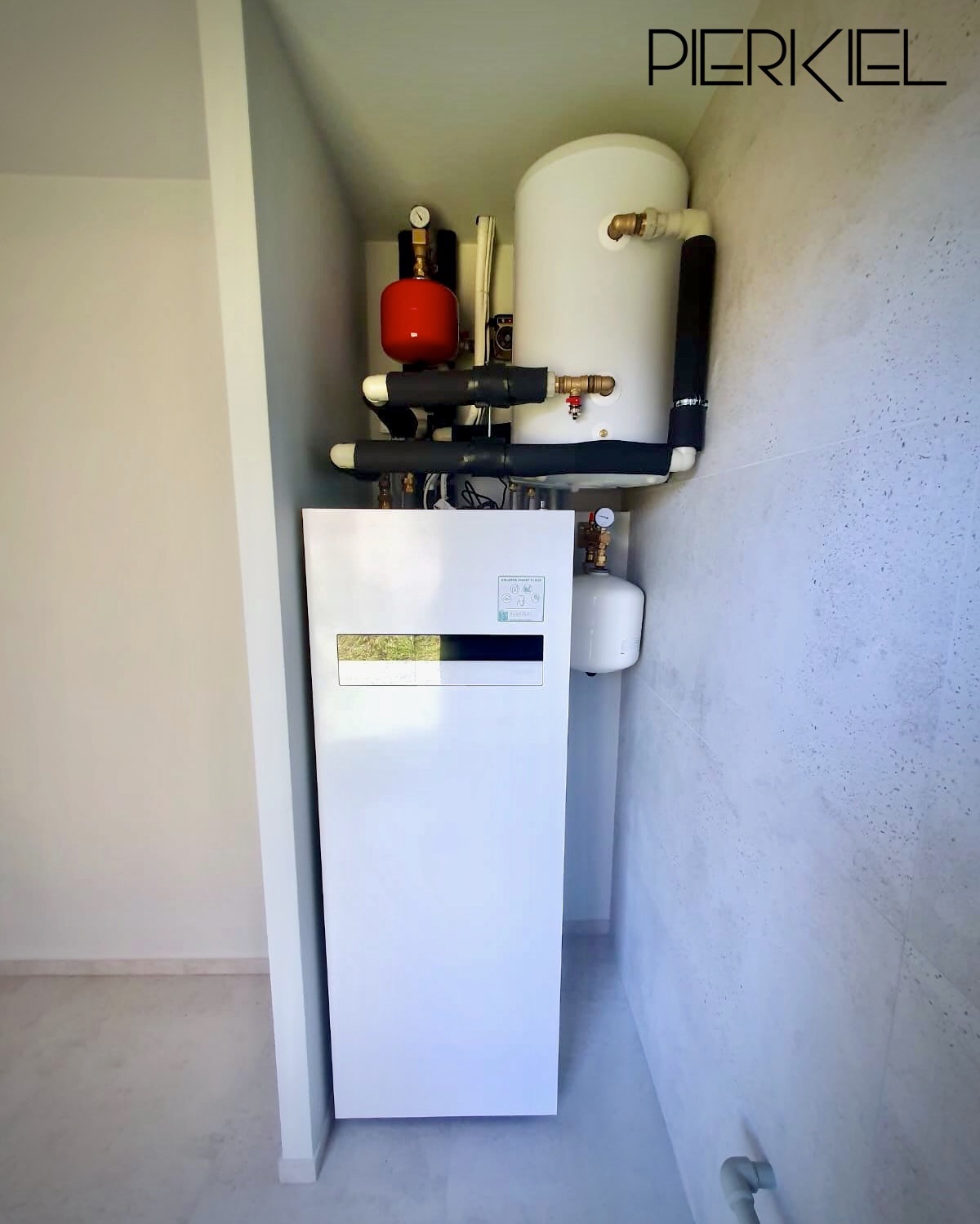 Biała pompa ciepła w kotłowni, z czerwonym zbiornikiem wyrównawczym i białym zasobnikiem ciepłej wody. Widoczne rury w izolacji i manometry. Minimalistyczne wnętrze.