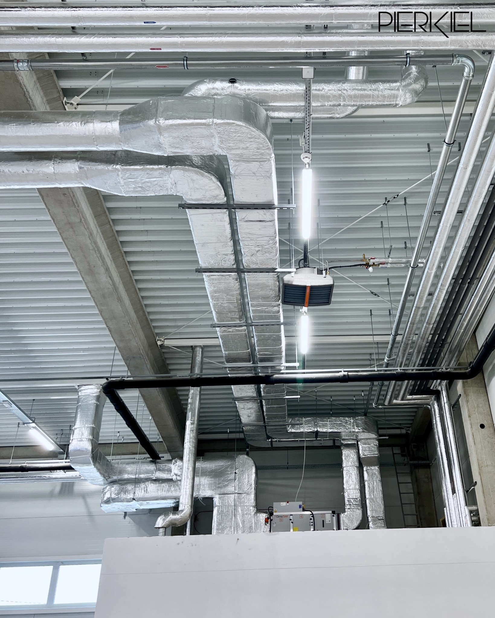 Instalacja wentylacyjna z srebrnymi, izolowanymi rurami pod sufitem hali. Widoczne elementy oświetlenia i konstrukcji budynku. Ujęcie z dołu.