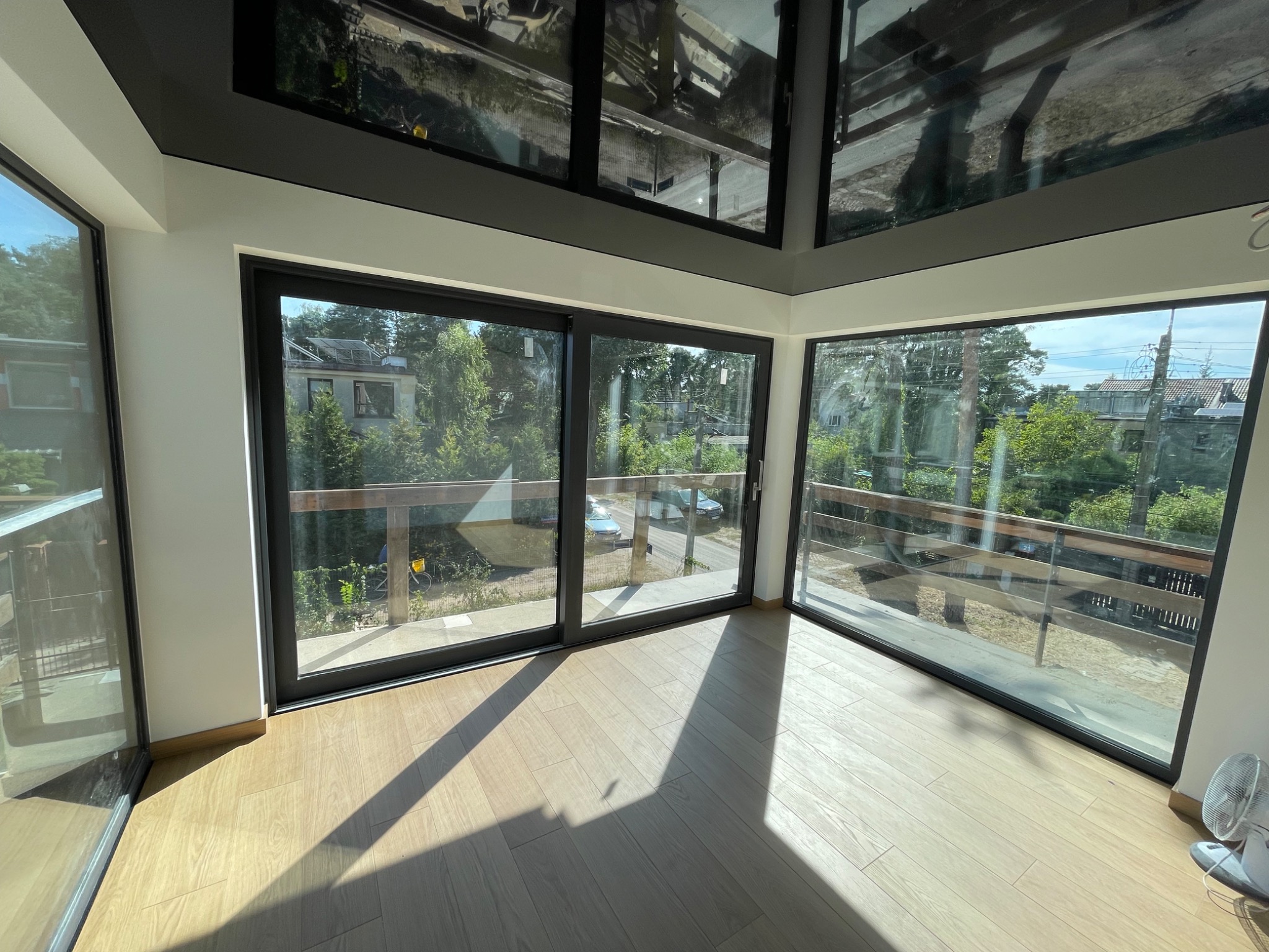 Jasne wnętrze z parkietem i dużymi oknami balkonowymi z czarnymi ramami. Widok na zieleń za oknem. Cień na podłodze. Nowoczesne wykończenie.