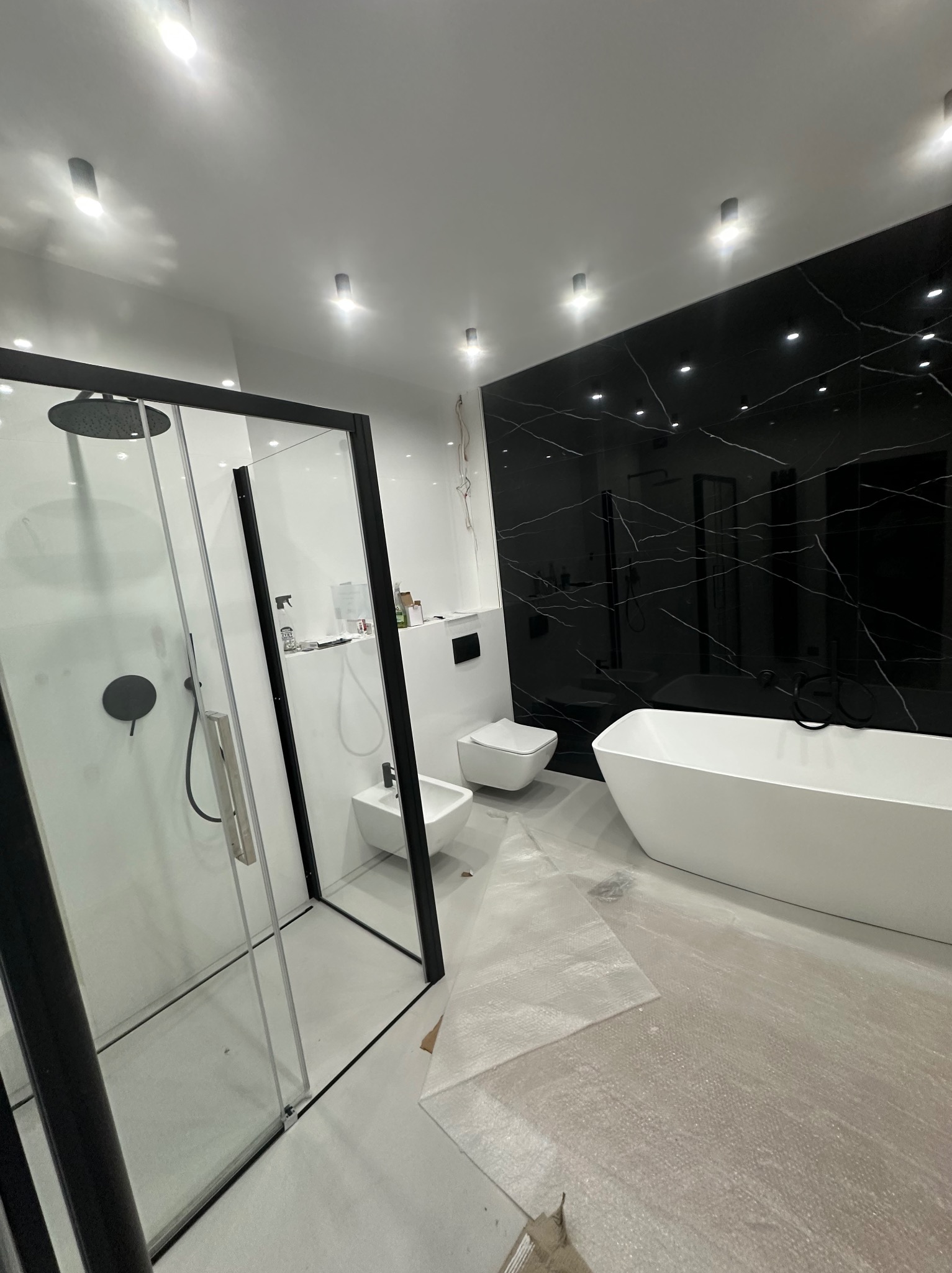 Nowoczesna łazienka z czarnymi płytkami imitującymi marmur, białą armaturą i kabiną prysznicową z czarnymi ramami. Podłoga zabezpieczona folią ochronną.