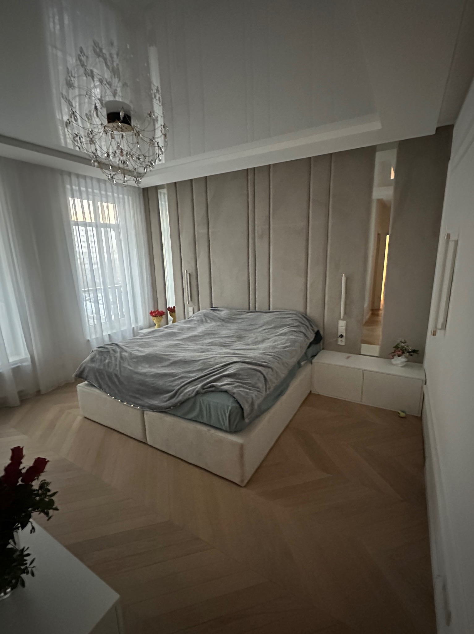 Elegancka sypialnia z łóżkiem kontynentalnym, panele ścienne w odcieniach beżu, parkiet w jodełkę i kryształowy żyrandol. Widok na okno z białymi zasłonami.